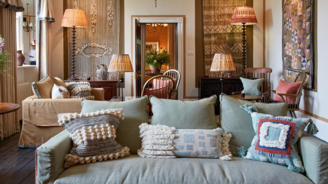 A cosy hotel lounge with a sage green sofa piled with textured cushions, rattan lampshades, and eclectic woven wall hangings in warm tones