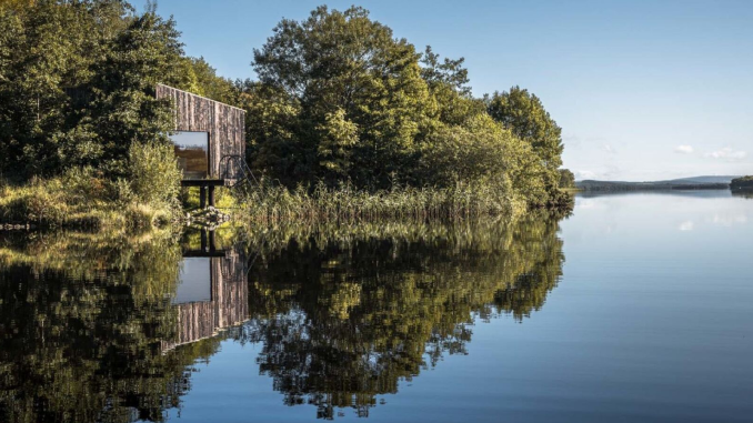 A weathered timber cabin perched at the edge of a glassy lough, perfectly reflected in the still water, surrounded by dense woodland under a clear blue sky