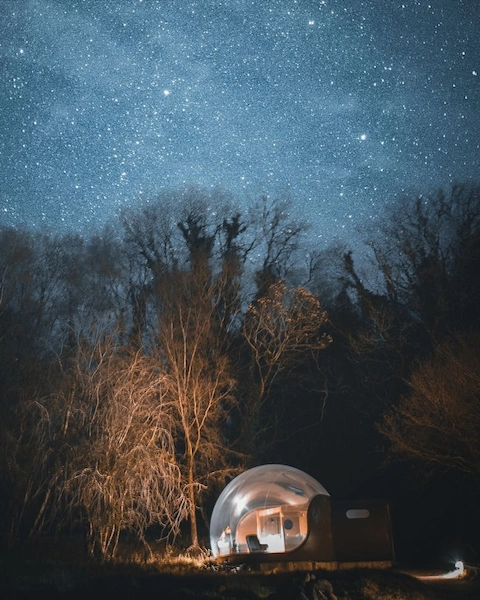 A glowing transparent bubble dome accommodation at Finn Lough, County Fermanagh, surrounded by bare winter trees beneath a vivid starlit night sky