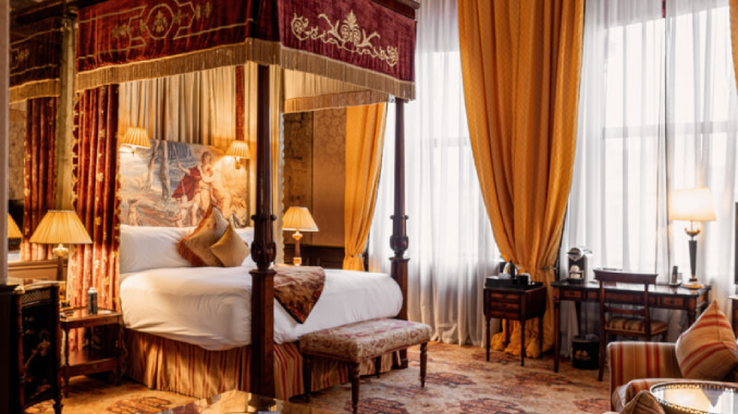 An opulent four-poster bedroom at the Merchant Hotel in Belfast, dressed in rich gold and burgundy fabrics, tapestry headboard, antique furnishings and floor-length drapes