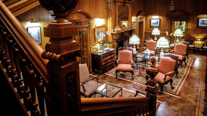 A grand oak-panelled hotel hall viewed from a carved staircase, with antique pink armchairs, Persian rugs, and a fireplace lit by Tiffany lamps