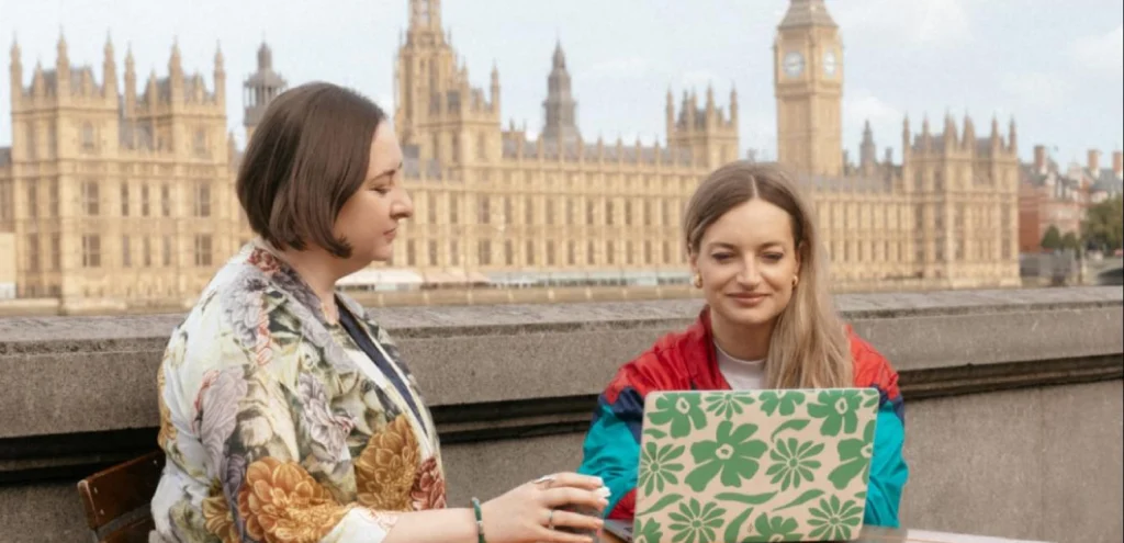 Two Tiernan Travel team members sitting at an outdoor table with a laptop, with the Houses of Parliament and Big Ben behind them in London