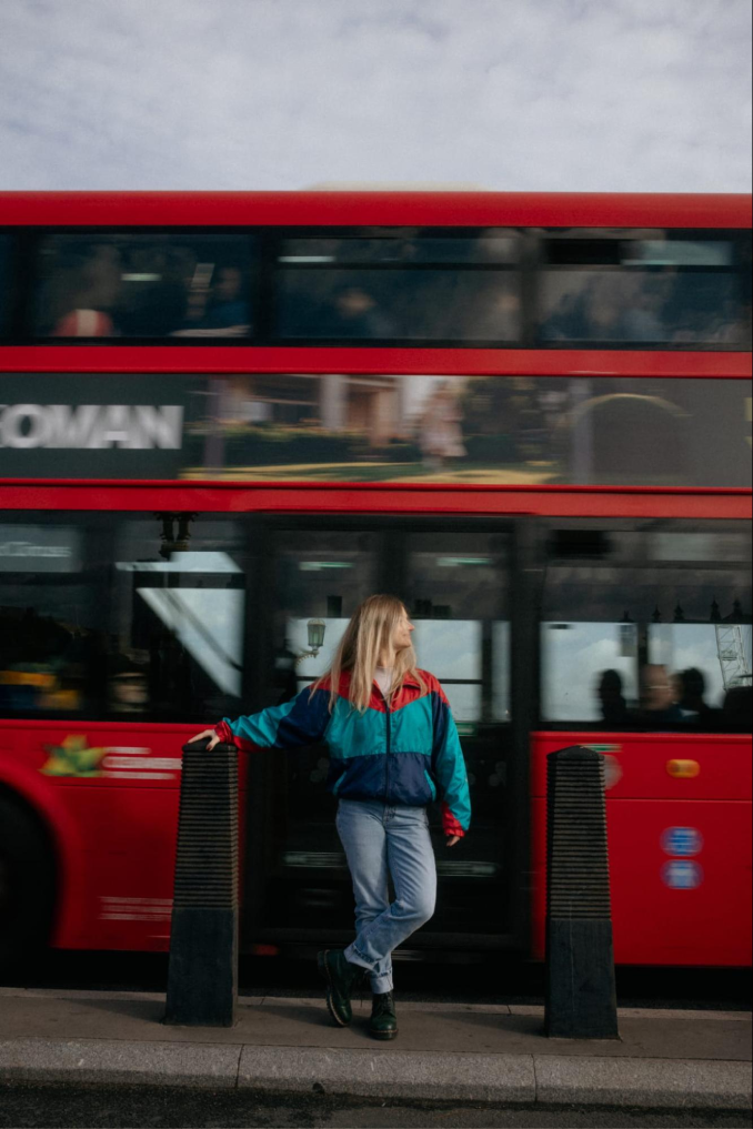 A Tiernan Travel team member in a colorful retro jacket leaning against a bollard as a red London double-decker bus passes behind her
