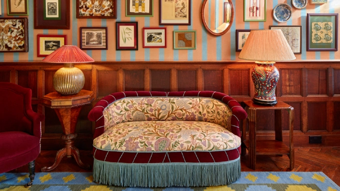A velvet canapé sofa with floral tapestry upholstery and teal fringe, flanked by ceramic lamps, beneath a gallery wall on striped wallpaper above wood panelling